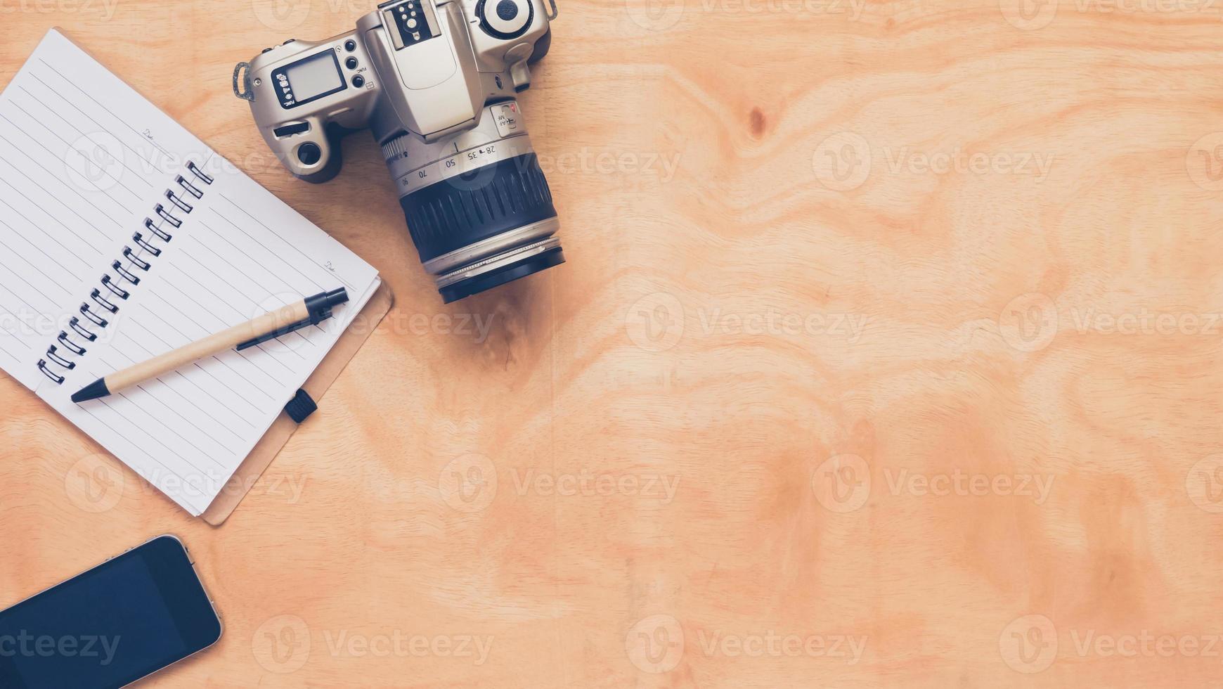 vista superior de la cámara con cuaderno, bolígrafo y teléfono móvil sobre fondo de mesa de madera. foto