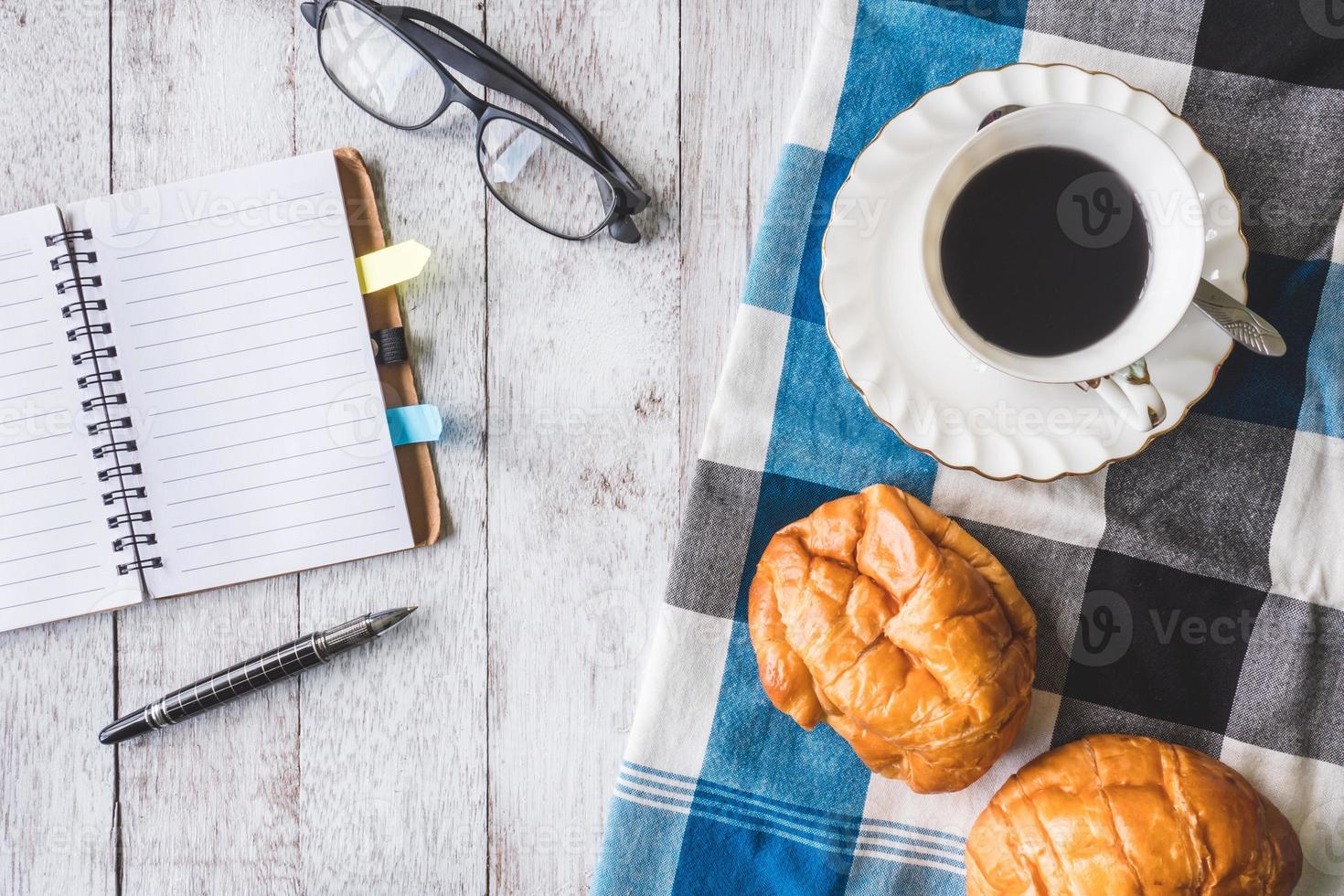 Top view of Coffee cup with bread, tablecloth, notebook, pen and glasses on wooden table background photo