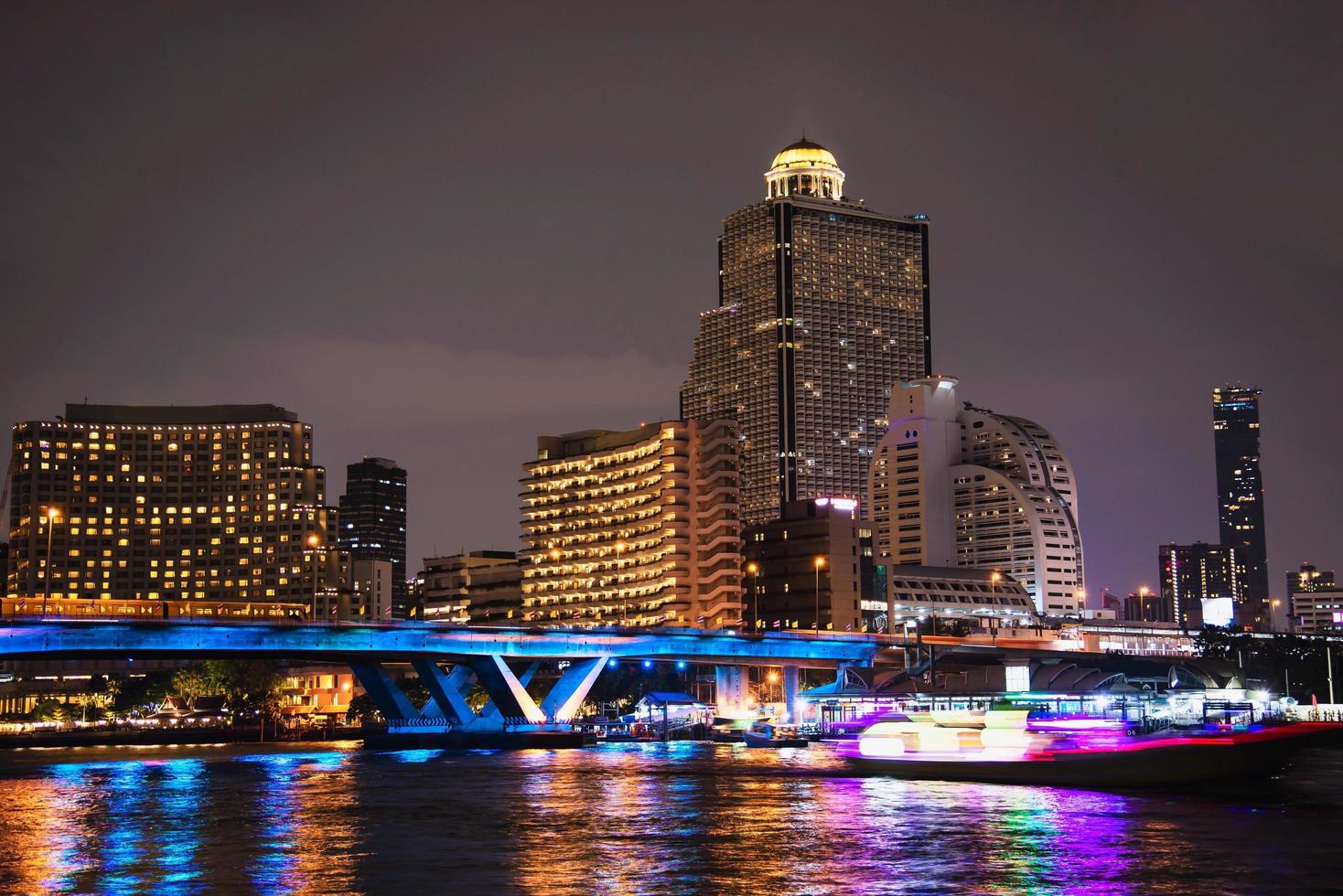 imagen nocturna colorida de la ciudad asiática - puente de luz azul sobre un gran río y fondo de construcción en bangkok, tailandia foto