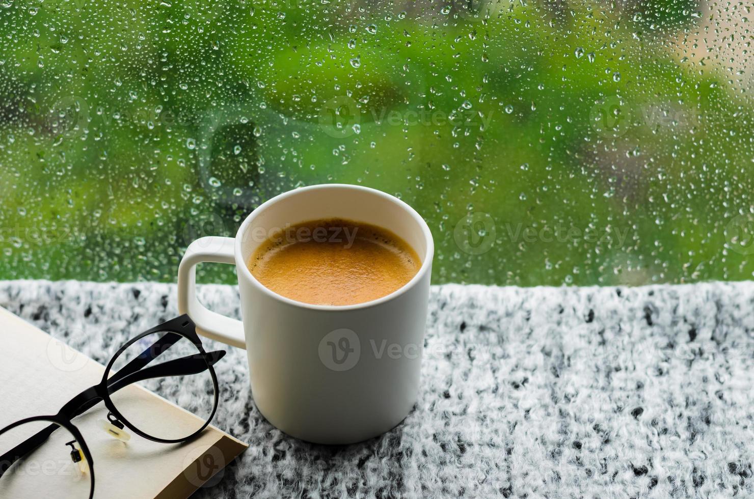 A cup of hot coffee with book and spectacles on table in morning with rain drop on window and green background. Stay home and relaxing concept. photo