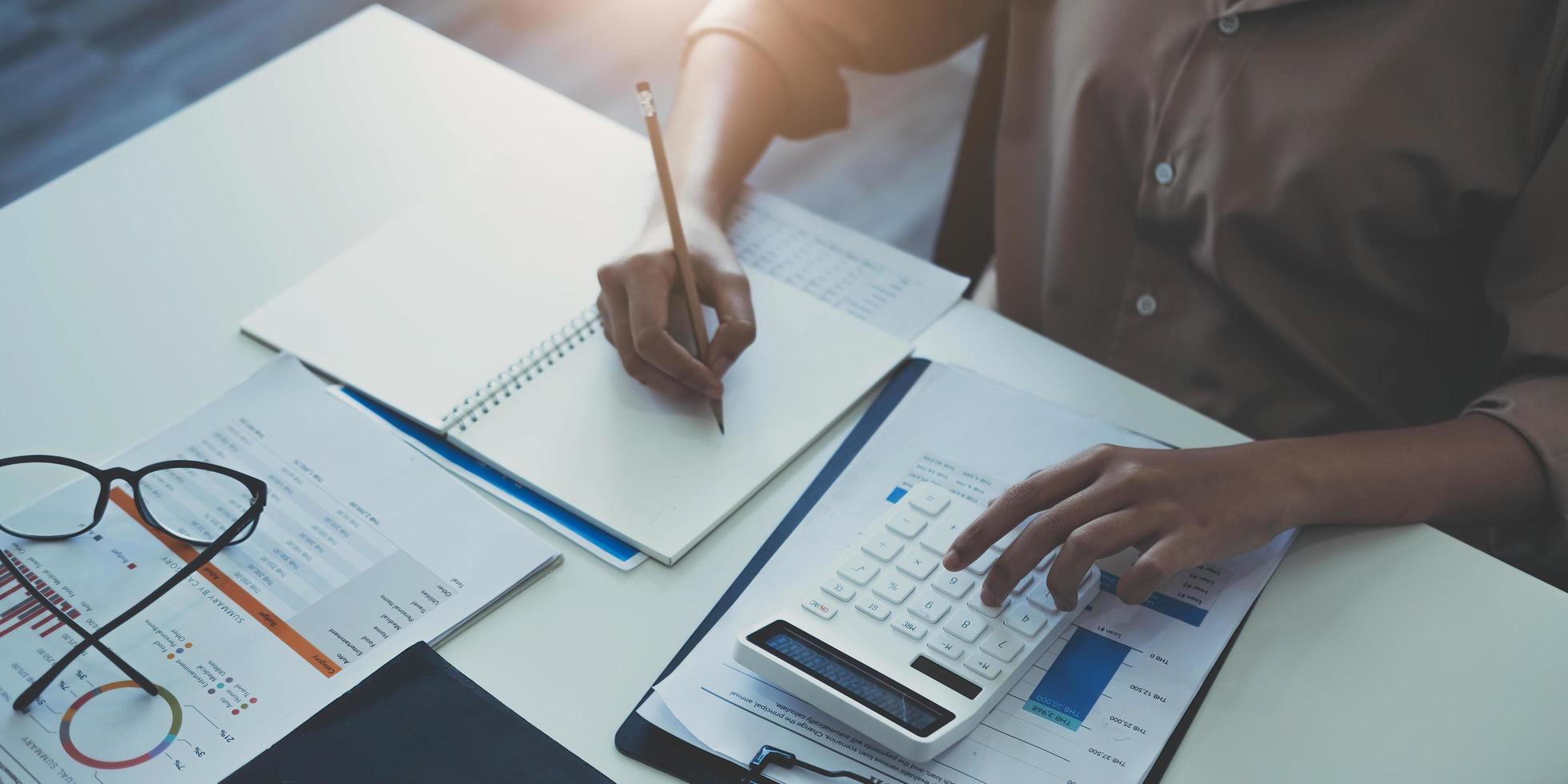 mujer de negocios usando calculadora y escribiendo nota con cálculo. mujer que trabaja en la oficina con computadora portátil y documentos en su escritorio foto