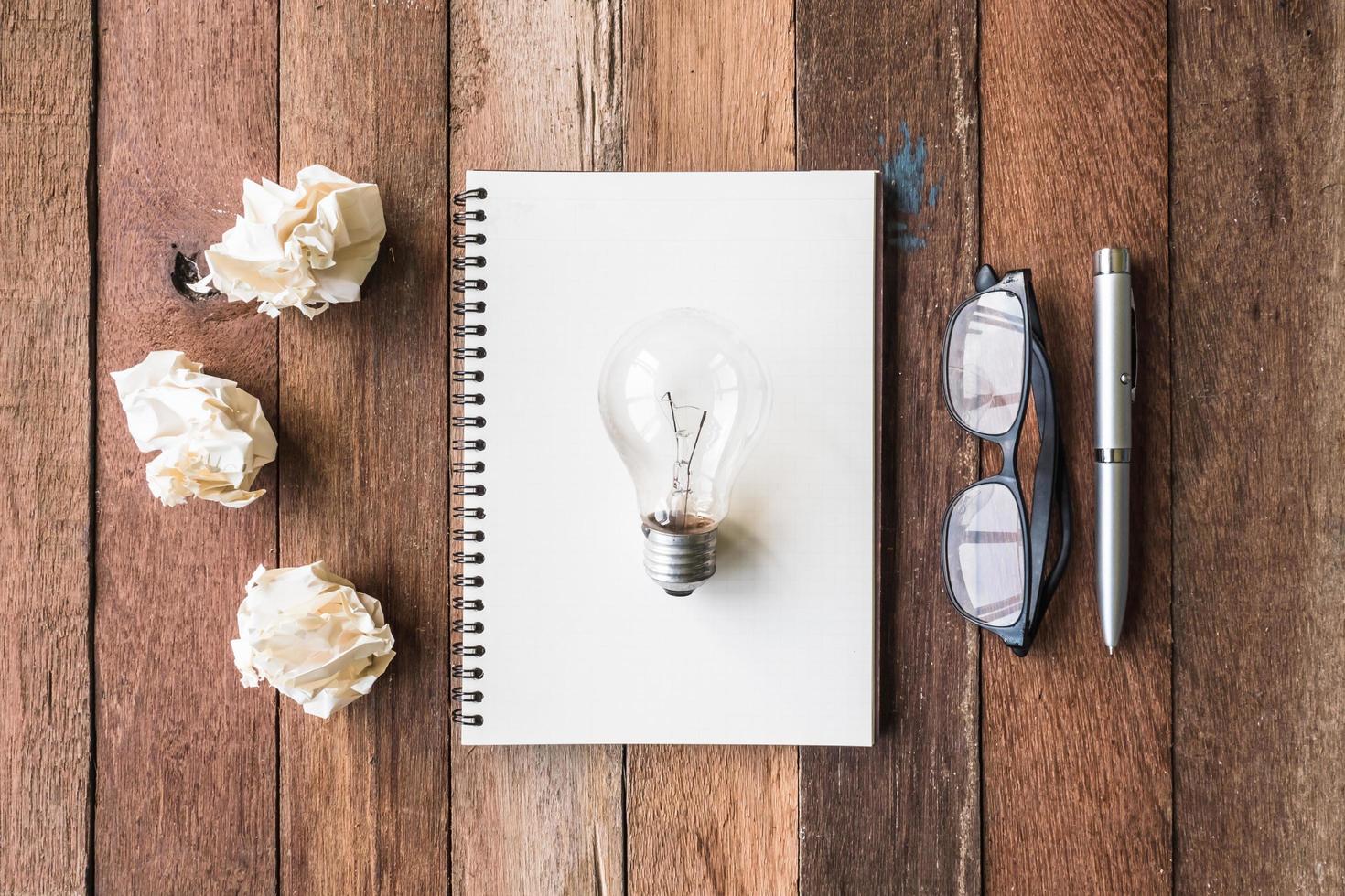 Top view of notebook with crumpled paper balls, pen, glasses and light bulb  on wooden table background. photo