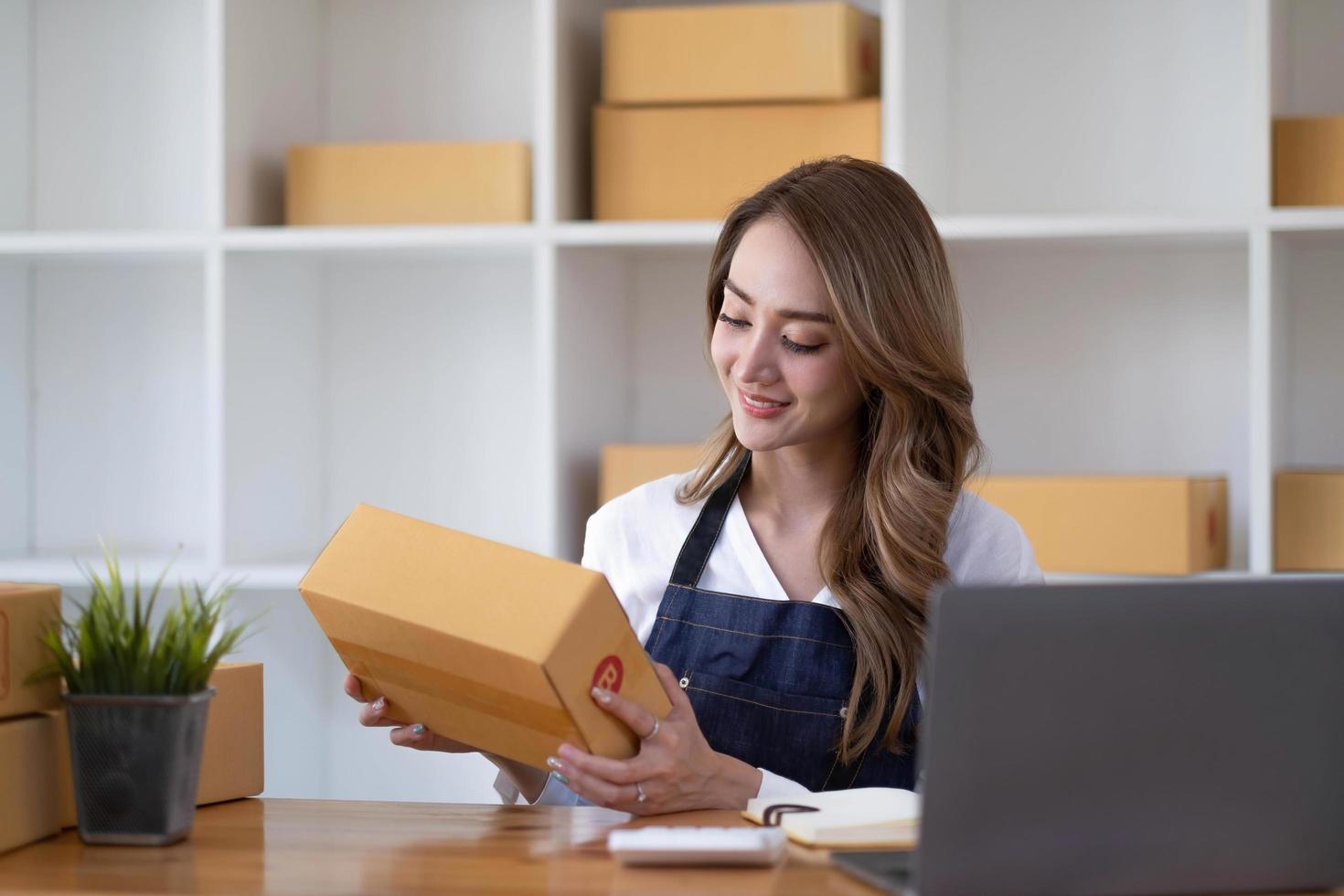 Young beautiful happy asian business woman owner of SME online using laptop receive order from customer with parcel box packaging at her startup home office, online business seller and delivery photo