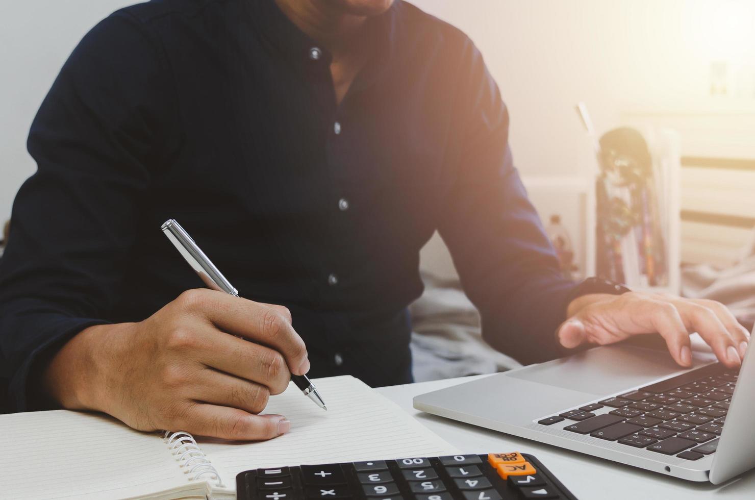 Businessman hand holding pen writing notebook and using keyboard computer laptop on desk. photo