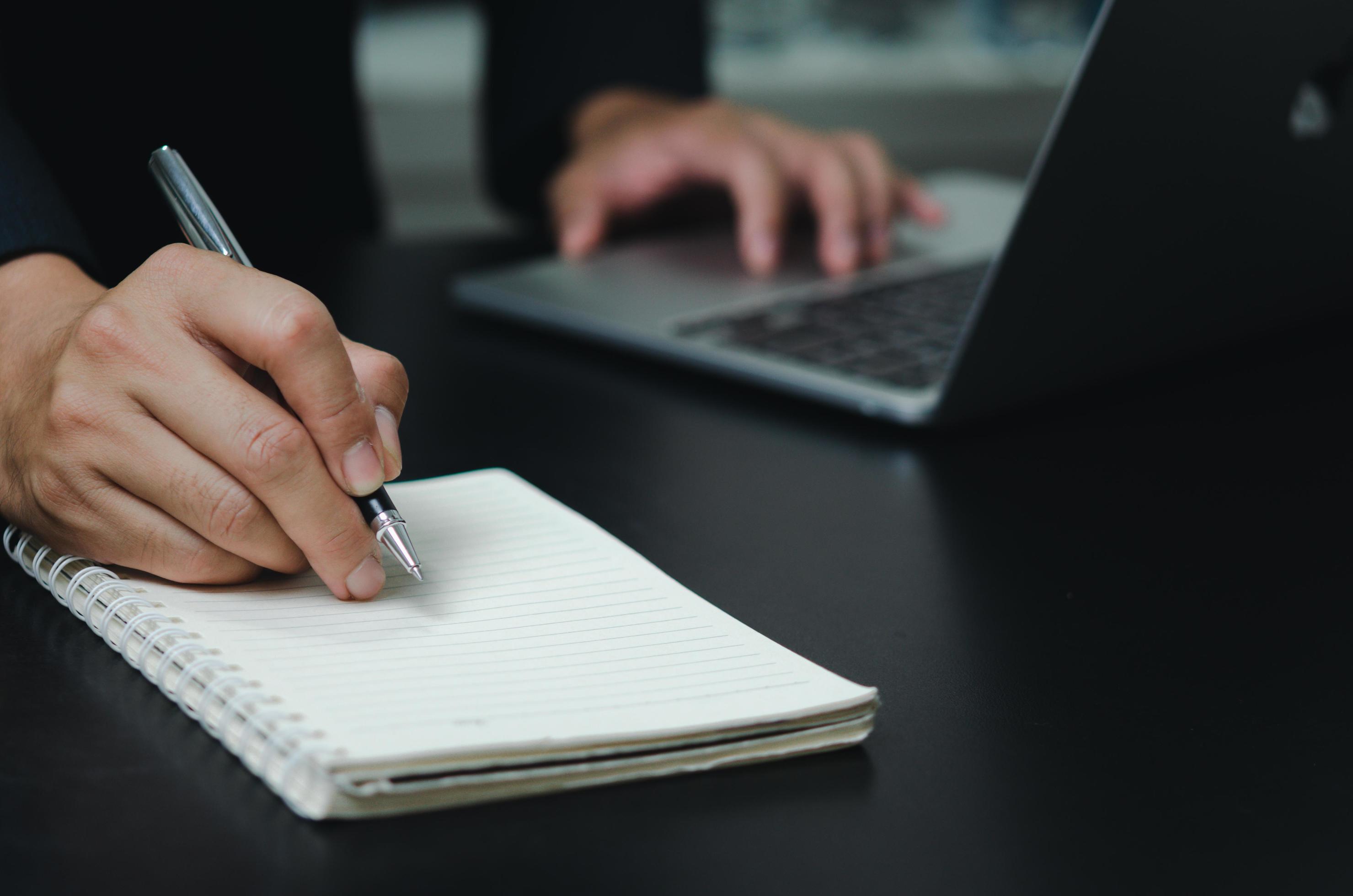 Businessman holding pen writing book note on desk with computer laptop