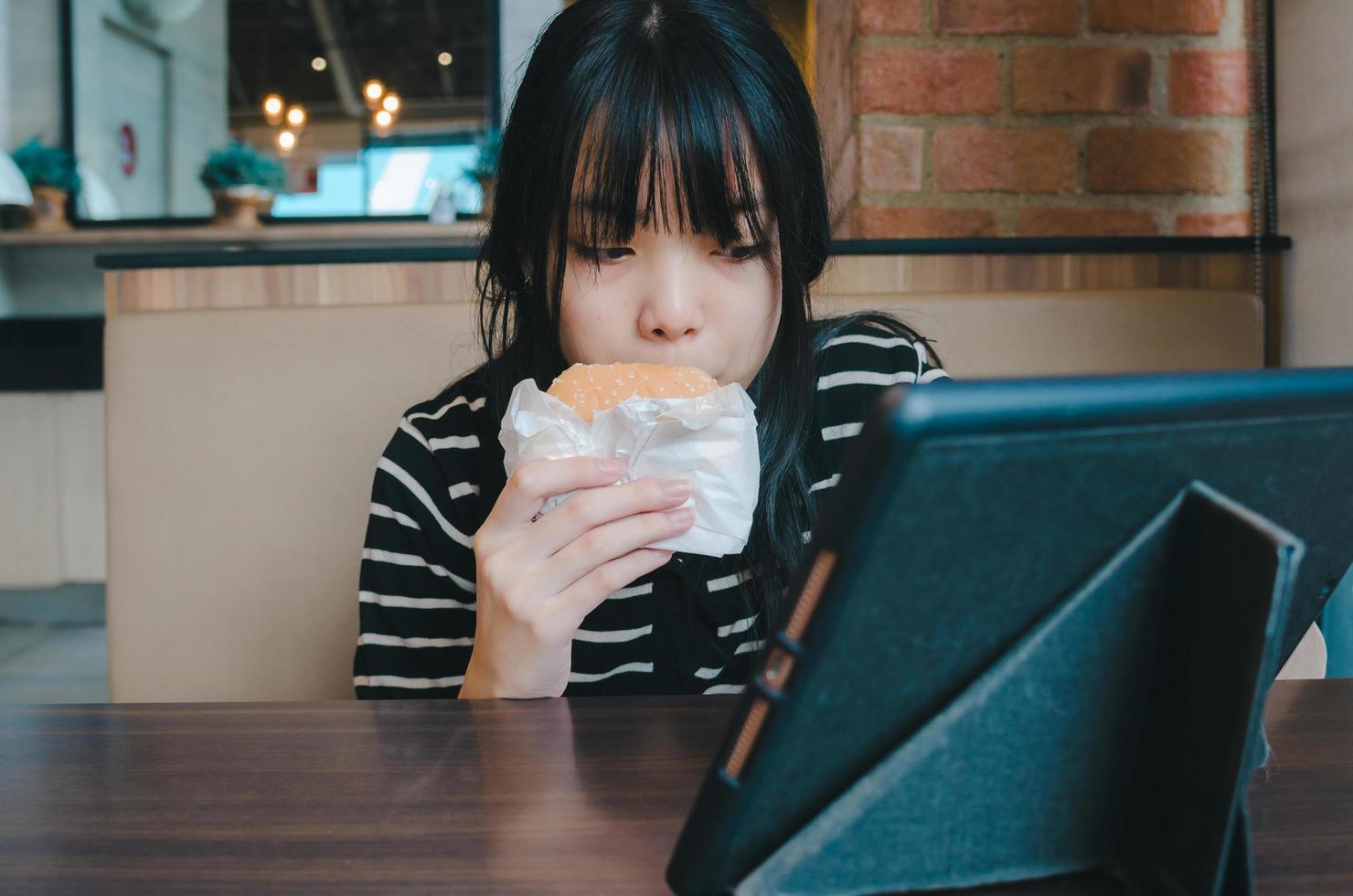 la joven está comiendo una hamburguesa y viendo una tableta tecnológica de medios sociales en línea sobre la mesa. foto