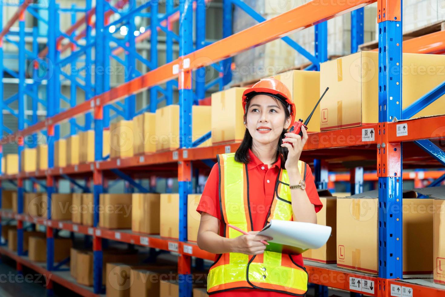 Warehouse logistic management business. young Asian woman wearing reflective safty vest and white helmet, Communicating via walky-talky in the warehouse factory as worker photo