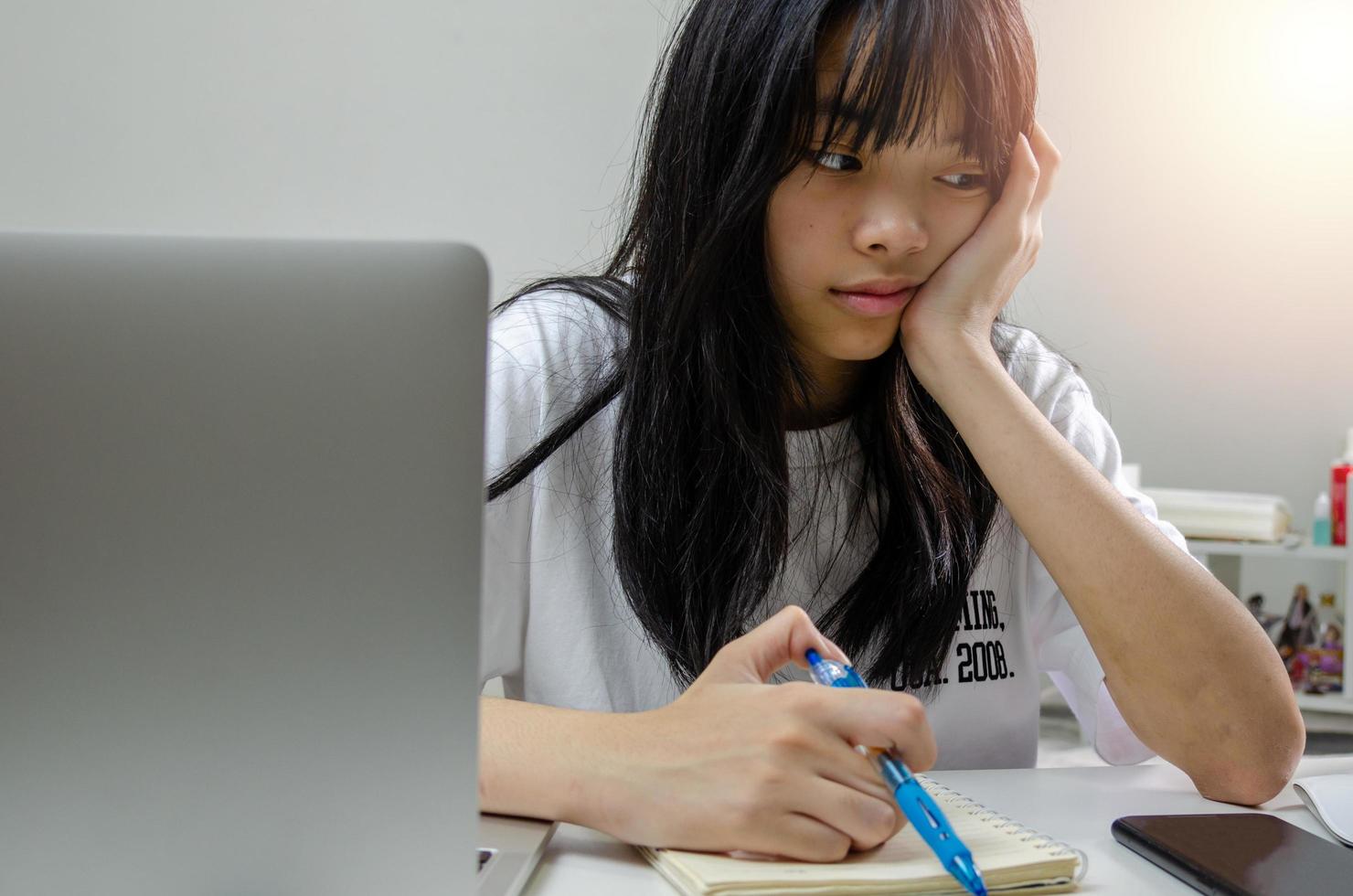 niña sosteniendo un bolígrafo escribiendo un libro de estudio aprendiendo un video digital de internet en línea computadora portátil en casa. foto
