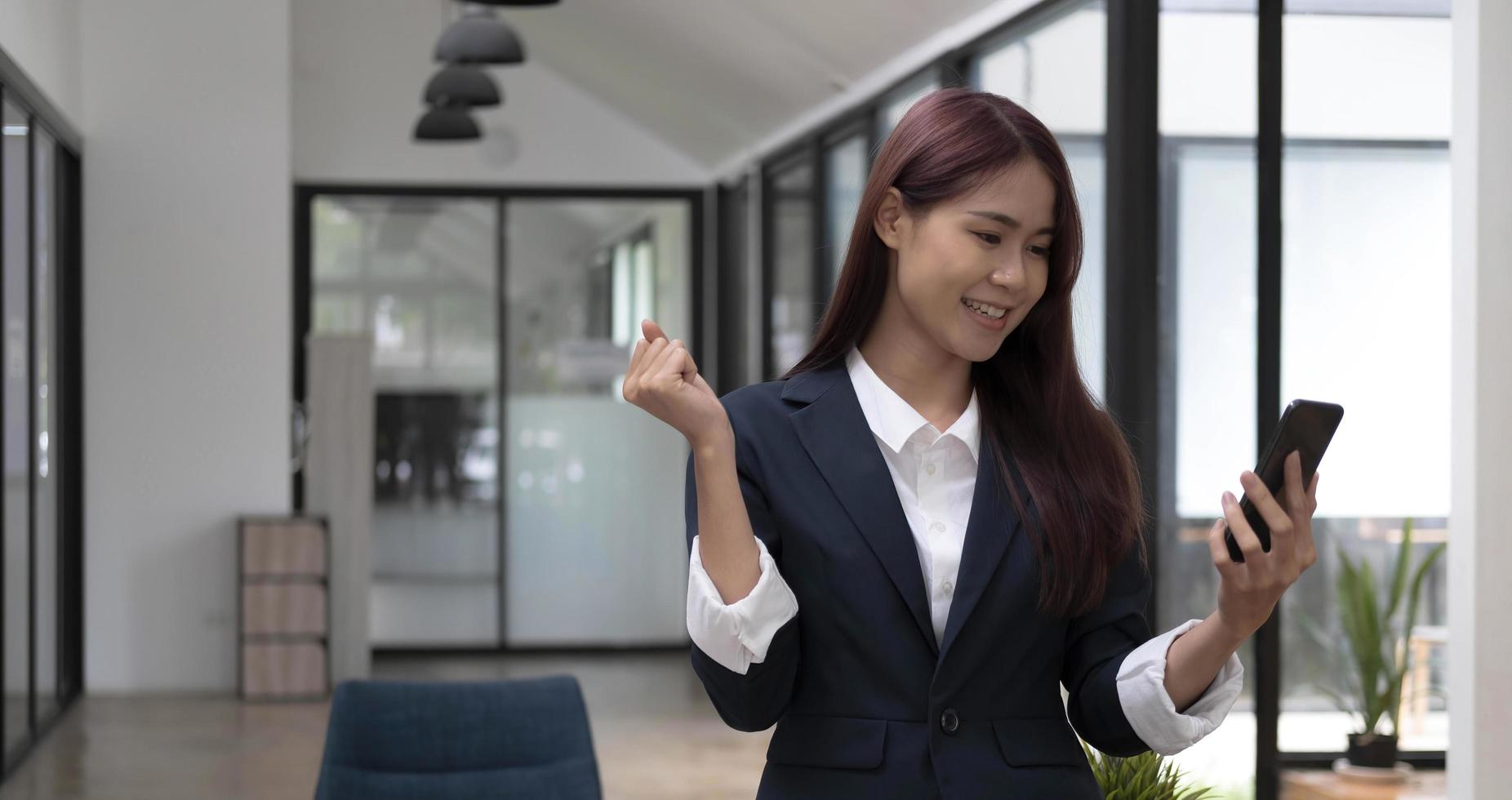 Cheerful amazed successful young Asian businesswoman standing in the office, using a smartphone, getting a surprised news. photo