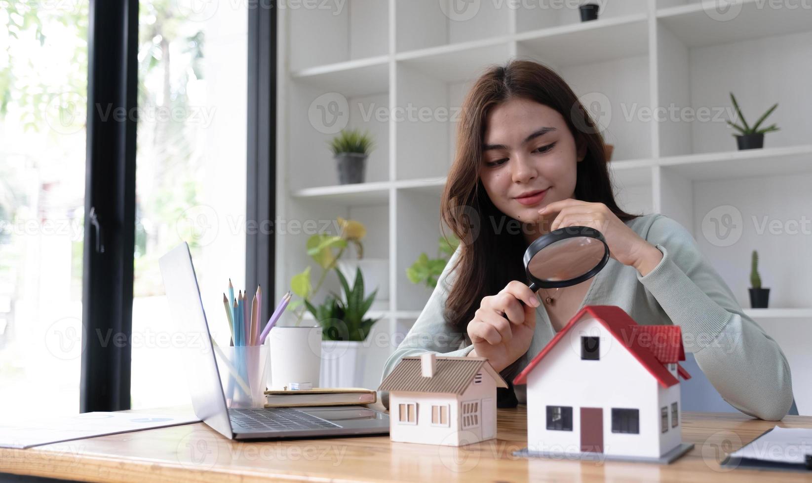 Happy woman hand holding magnifying glass and looking at house model, house selection, real estate concept. photo