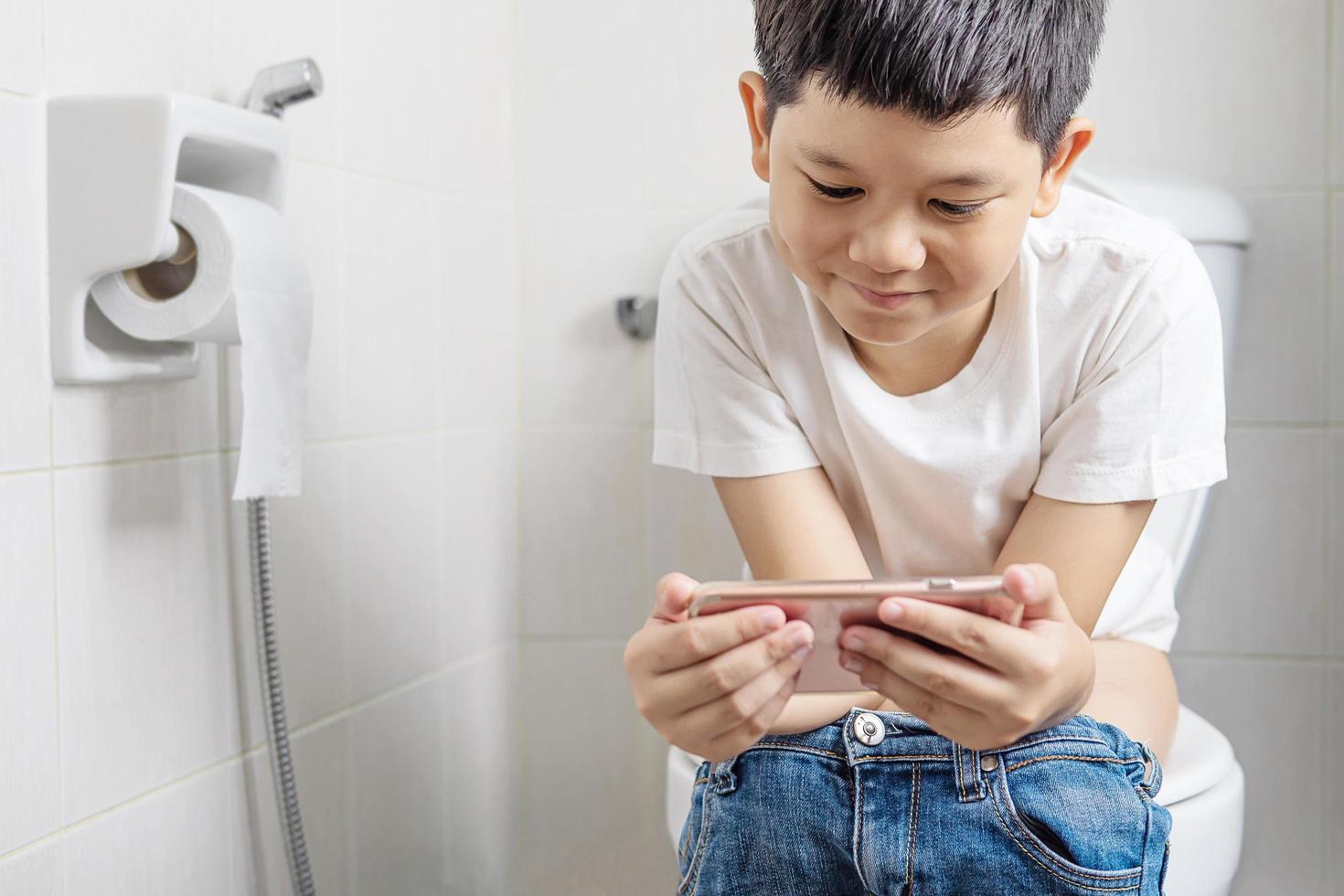 Asian boy sitting on toilet bowl while playing mobile phone - health problem concept photo