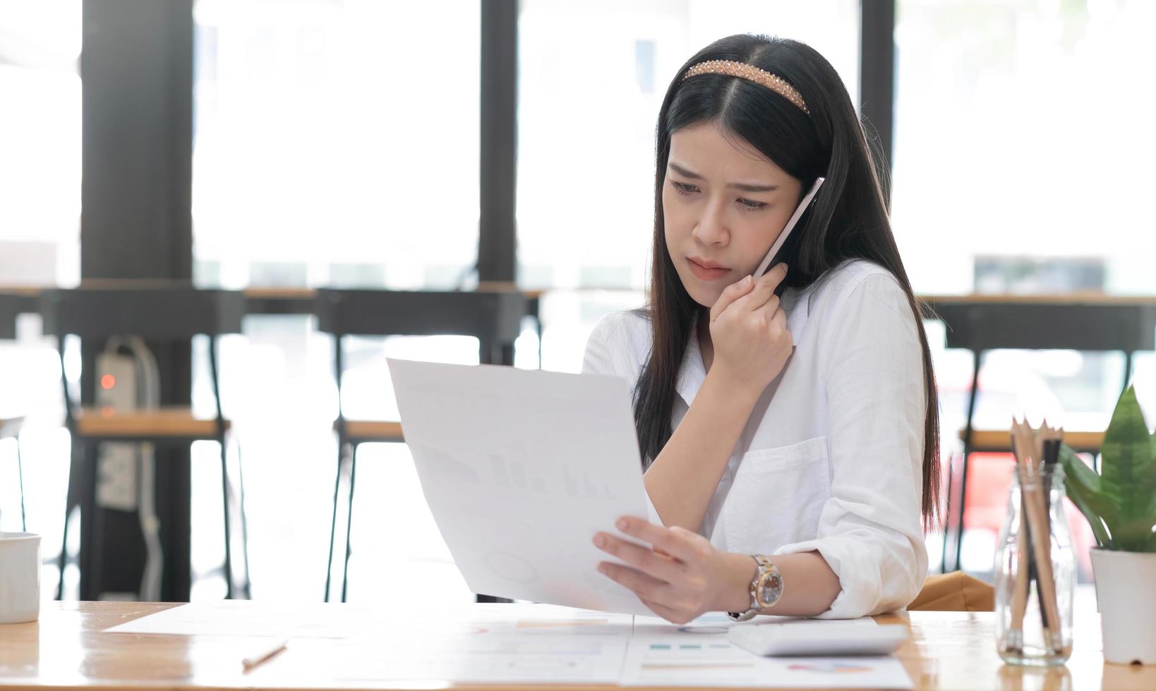 Serious and busy millennial Asian businesswoman or female marketing manager looking some details on paper while having a phone call with her partner. photo