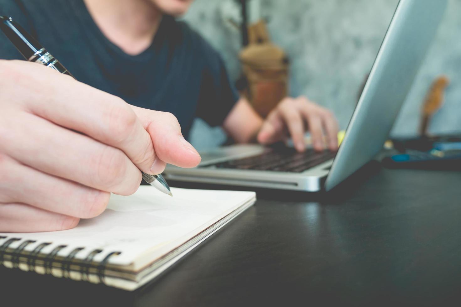 Casual young man writing some data in paper and using laptop on the desk photo
