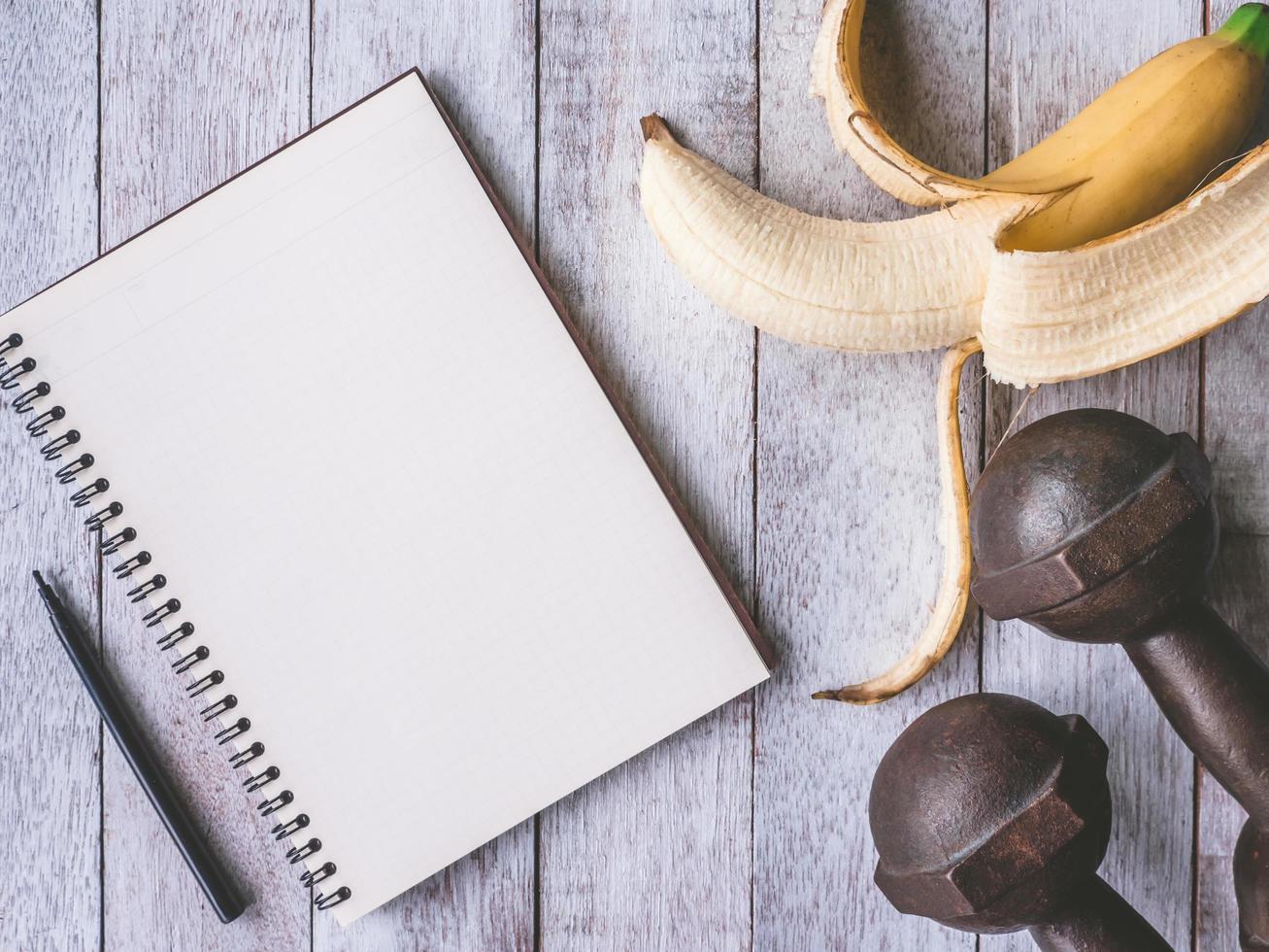 plátano con pesas de hierro y cuaderno en el fondo de la mesa de madera. concepto de entrenamiento y dieta foto