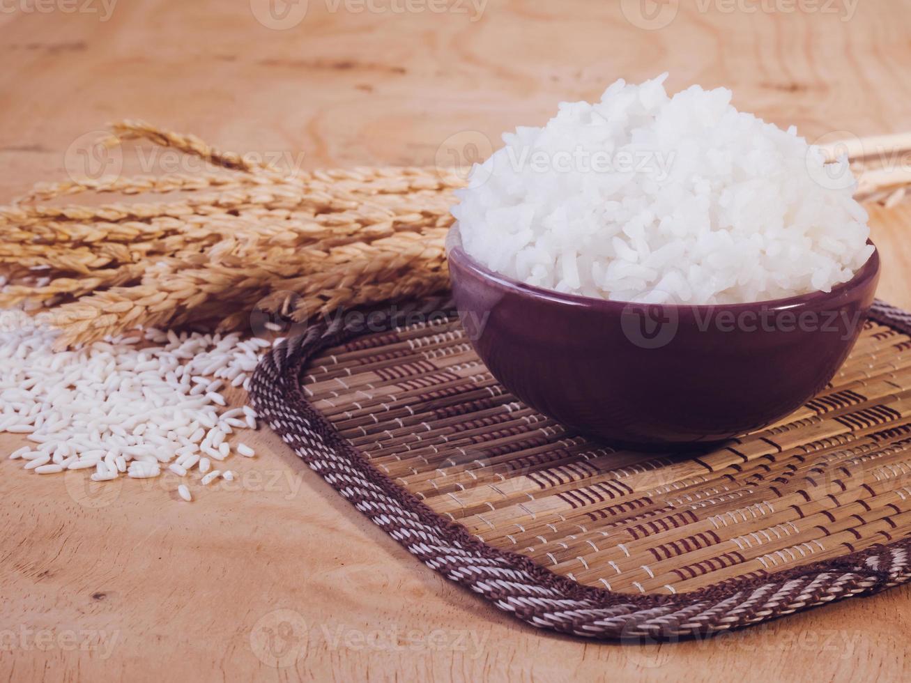 Cooked rice in bowl with raw rice grain and dry rice plant on  wooden table background. photo