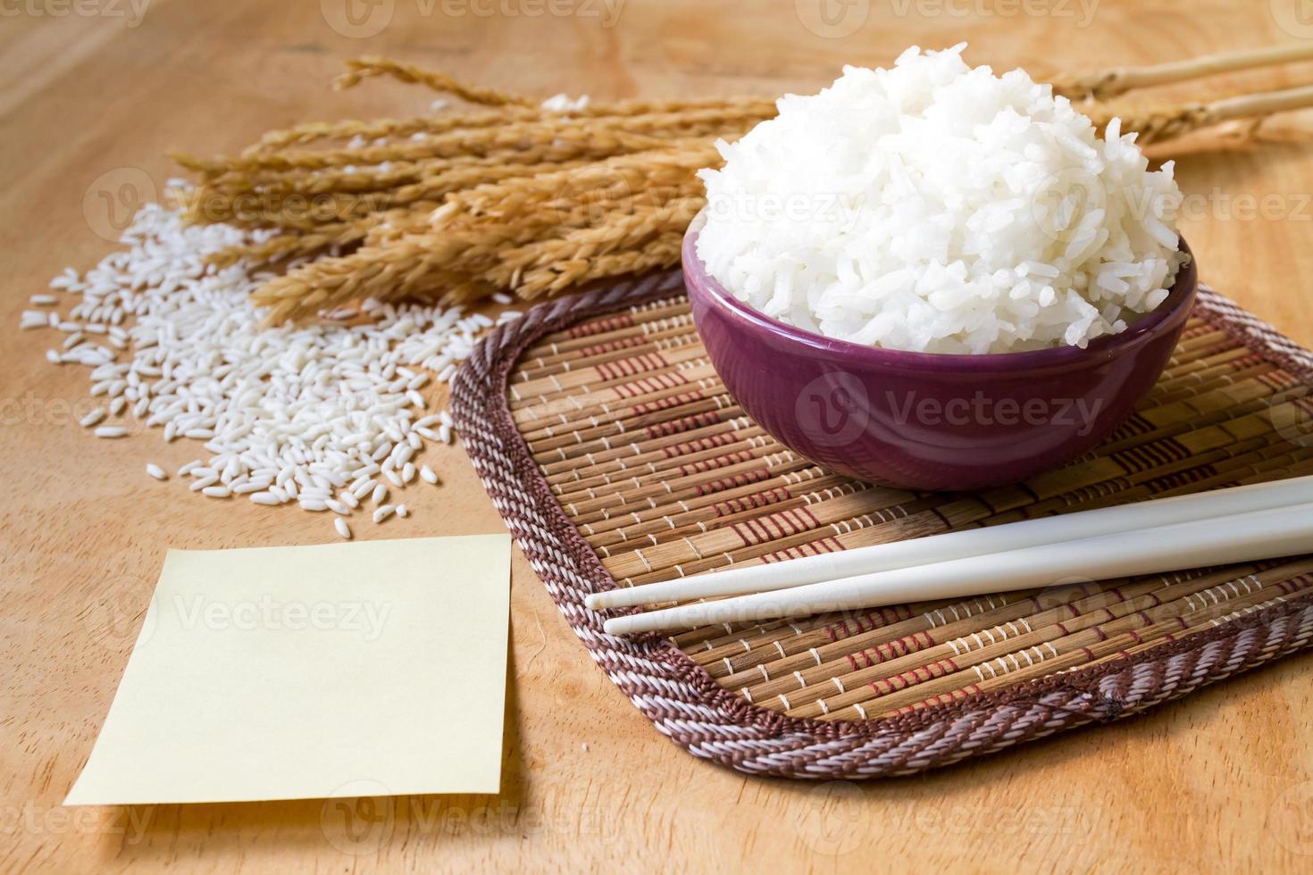 Cooked rice in bowl with raw rice grain ,dry rice plant and empty paper on  wooden table background. photo