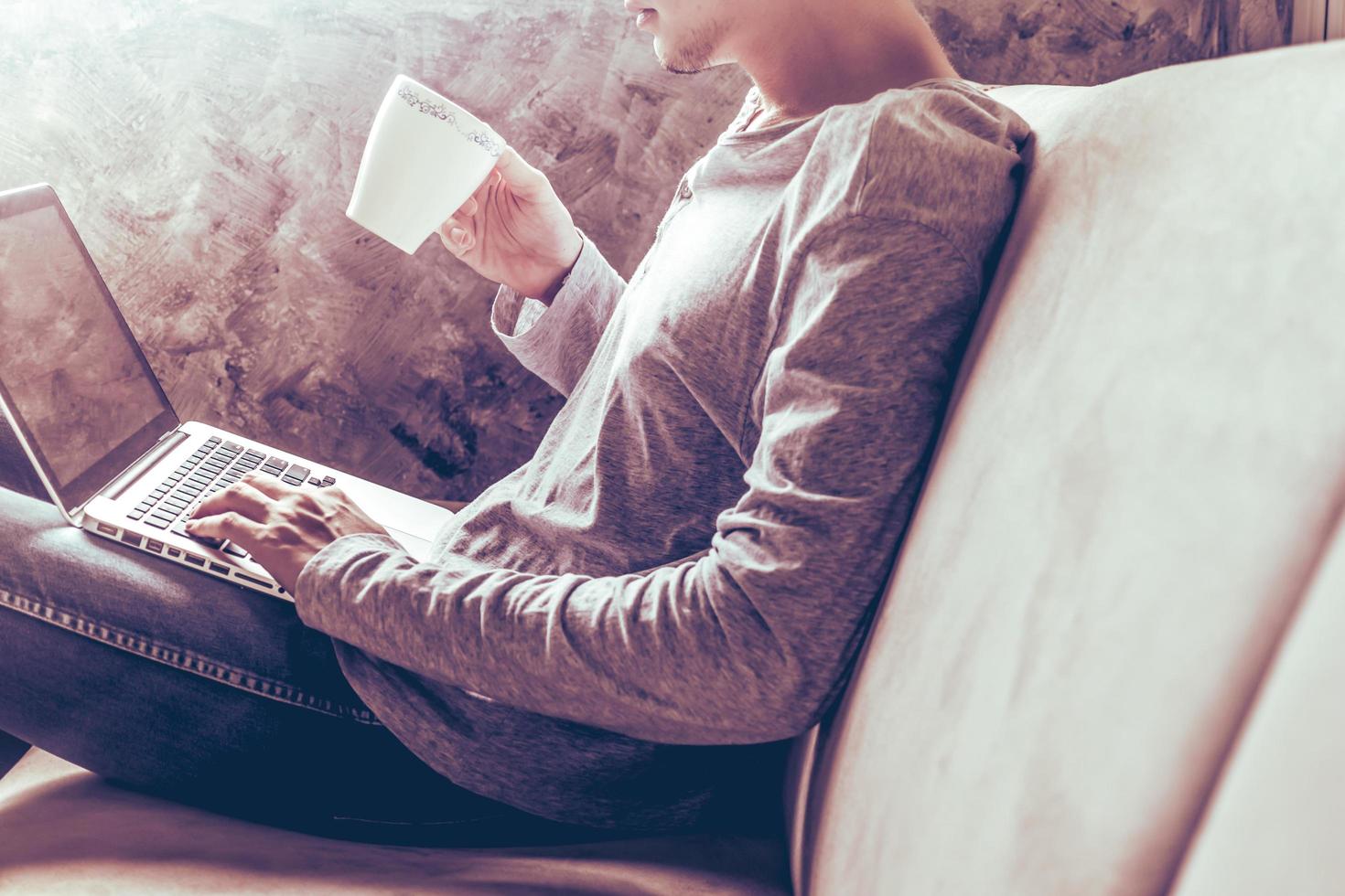 Casual young man using laptop and holding a cup of coffee on sofa at home. photo