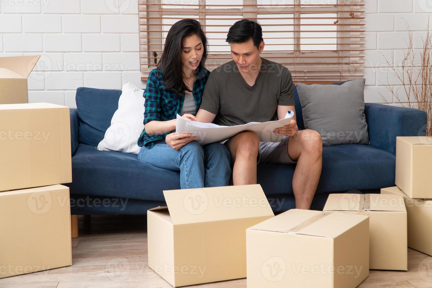 Smiling young Asian happy couple hold blueprint for home decoration ideas at moving day in their new home after buying real estate. Concept of starting a new life for a newly married couple. photo