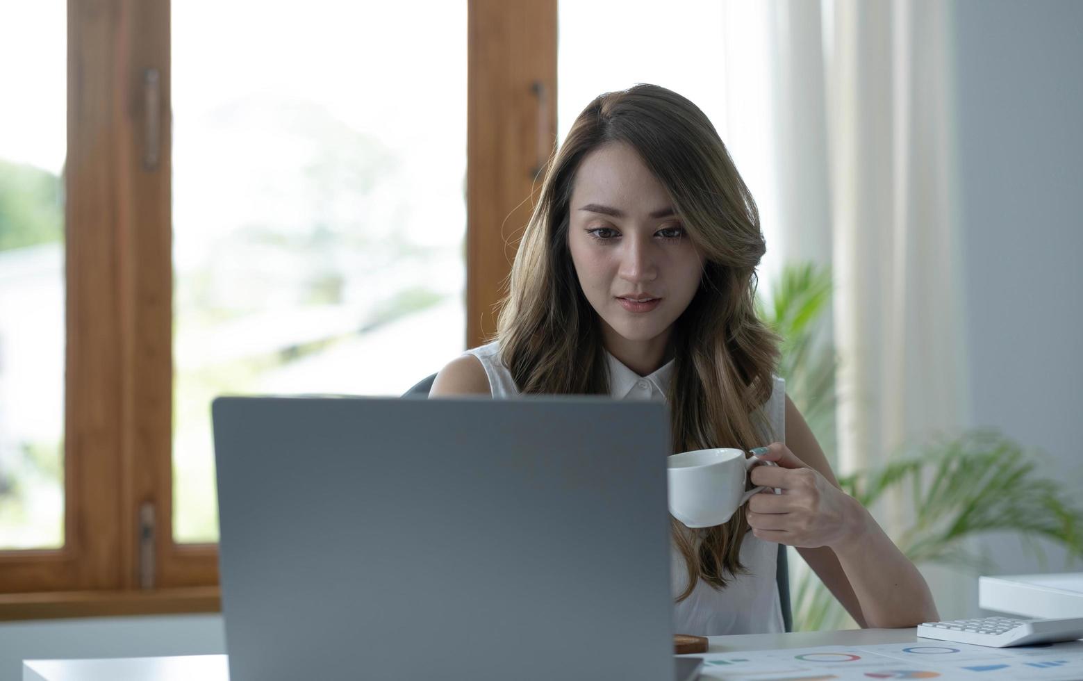 Photo of positive Asian woman holds a cup of coffee imagines plan holiday in office.