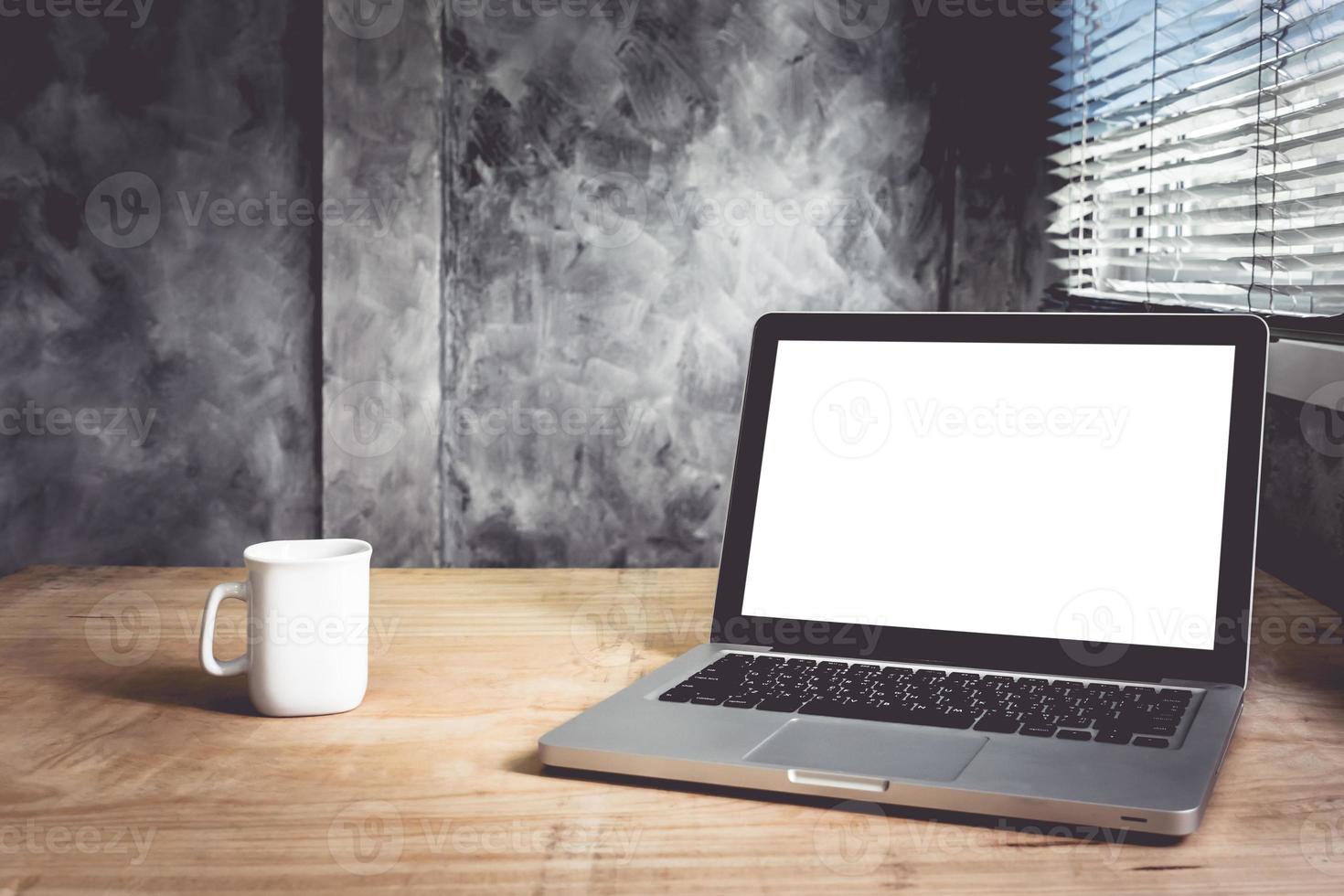 Laptop with white blank screen and cup of coffee on the wooden desk with grunge wall background. photo