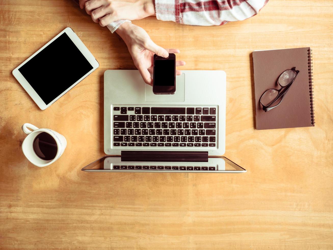 vista superior del hombre usando un teléfono inteligente con una computadora portátil y una tableta en el fondo de la mesa de madera. foto