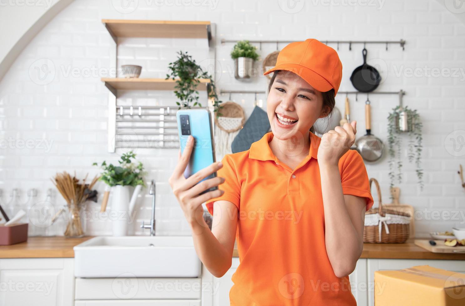 hermosa repartidora asiática con uniforme naranja envía productos de paquete para cocinar a los clientes en la cocina a través de un teléfono inteligente. transporte y logística nacional e internacional foto