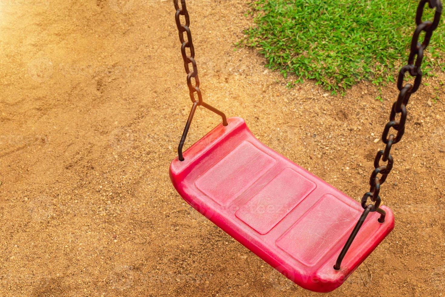 A red swing is attached to two old rusted chains in the playground. in the park saw the ground and the grass photo