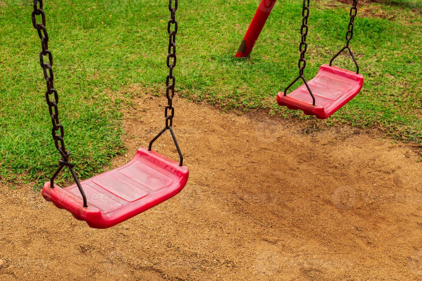 A red swing is attached to two old rusted chains in the playground. in the park saw the ground and the grass photo