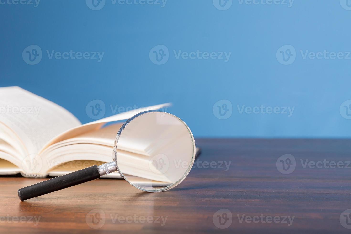 open book with magnifying glass on wooden desk in information library of school or university, concept for education,reading , study, copy space and blue background. photo