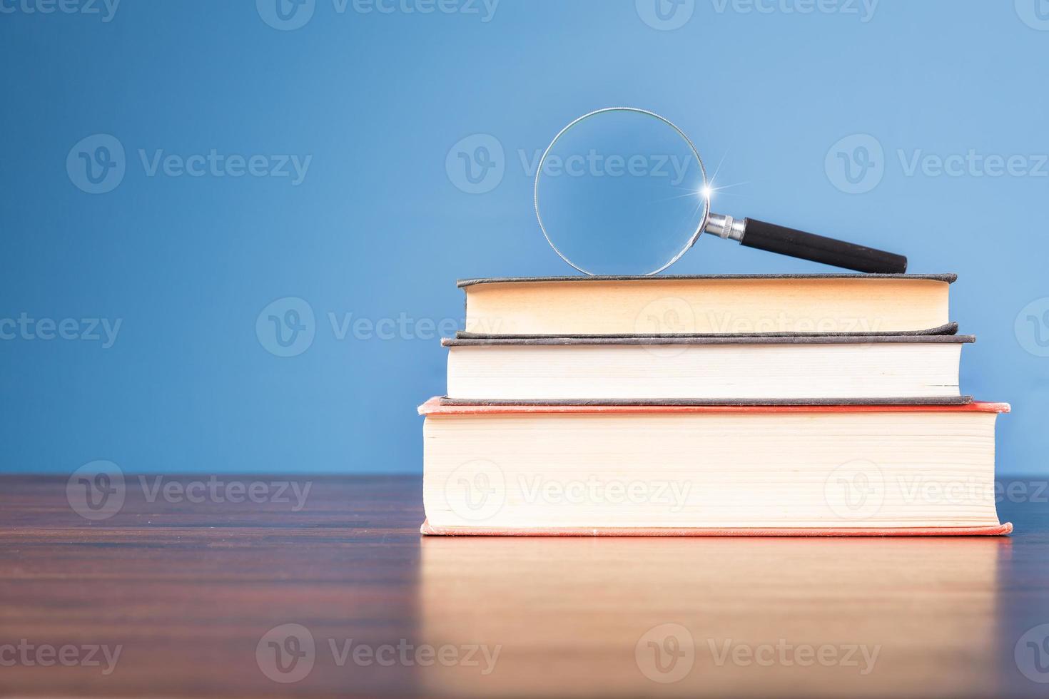 Stack book with magnifying glass on wooden desk in information library of school or university, concept for education, reading, study, copy space on blue background. photo