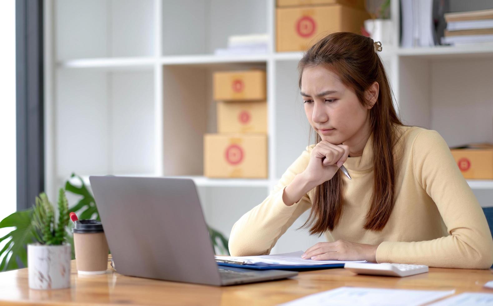 un retrato de una joven asiática seria que trabaja con una computadora portátil en la oficina llena de paquetes y cajas apiladas, una mesa ocupada, para sme, entrega, puesta en marcha de negocios y concepto de oficina en el hogar. foto