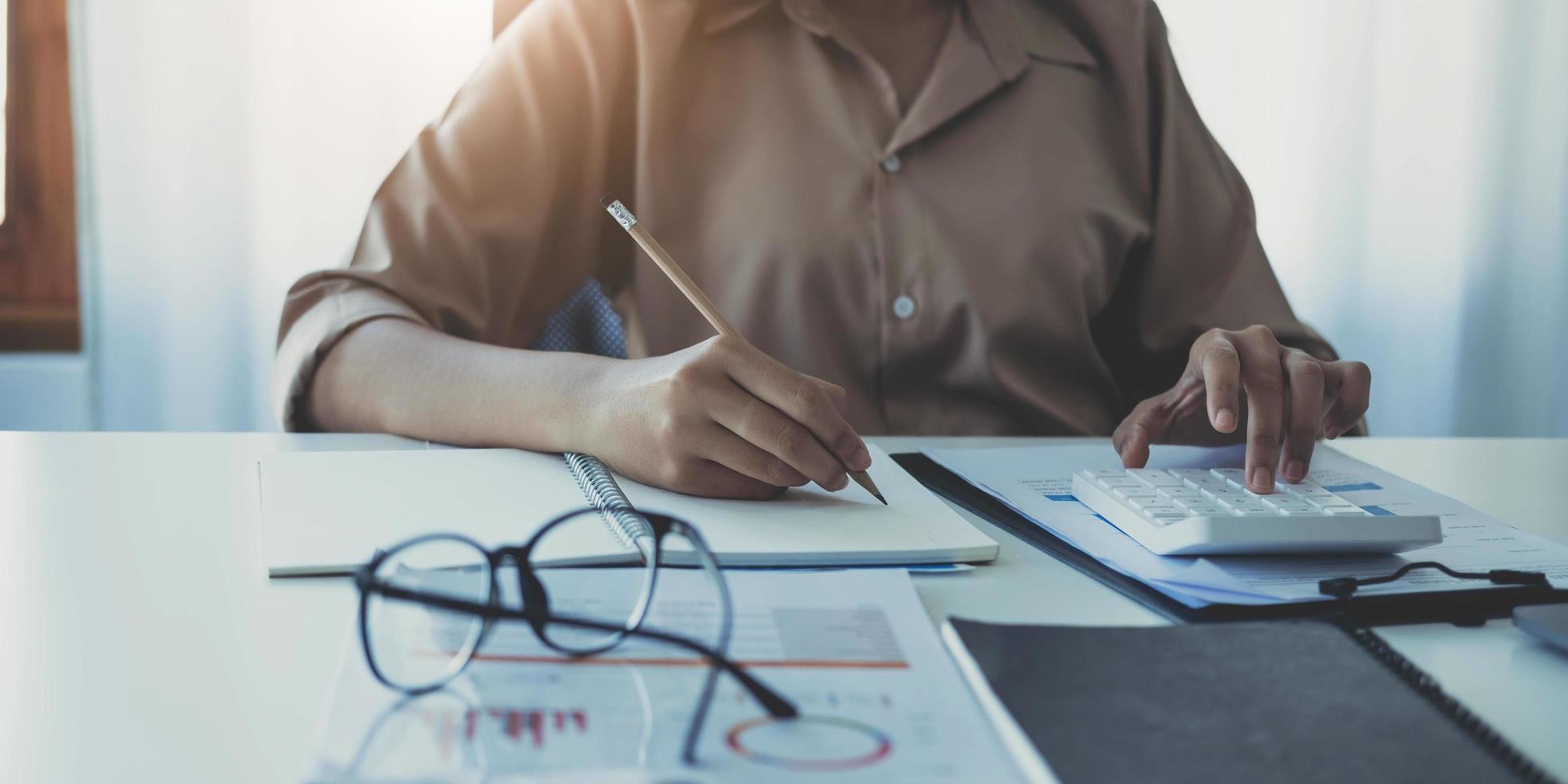 mujer de negocios usando calculadora y escribiendo nota con cálculo. mujer que trabaja en la oficina con computadora portátil y documentos en su escritorio foto