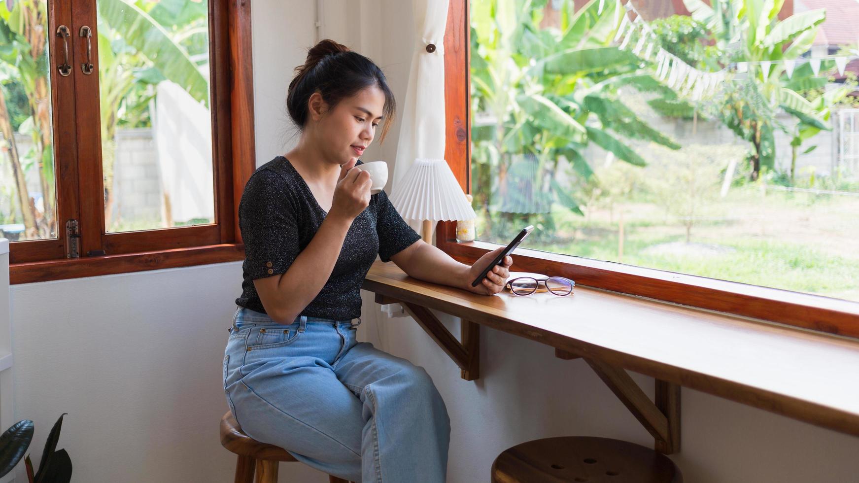 Asian woman with a beautiful smile watching on mobile phone during rest in a coffee shop, happy Thailand female sit at wooden bar counter drinking coffee relaxing in cafe during free time photo