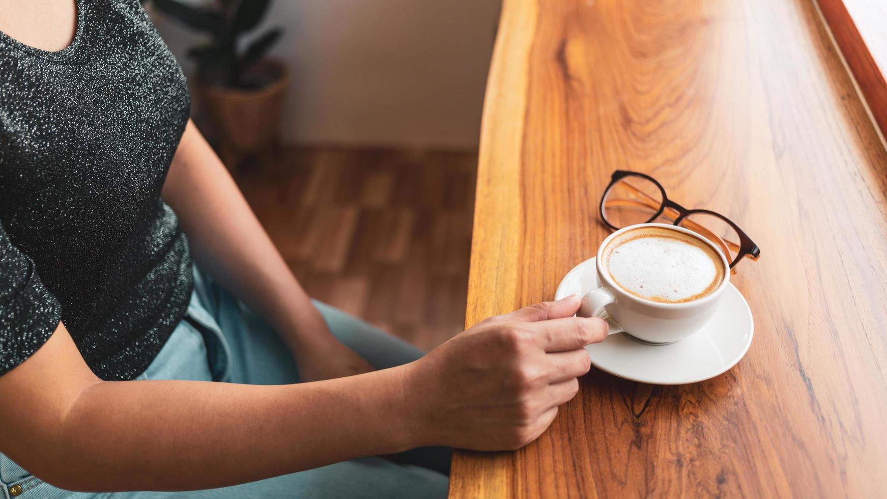 Beautiful Asian woman sits down at the bar counter inside a window coffee shop holding a cup of coffee, smiling relaxed in a cafe photo