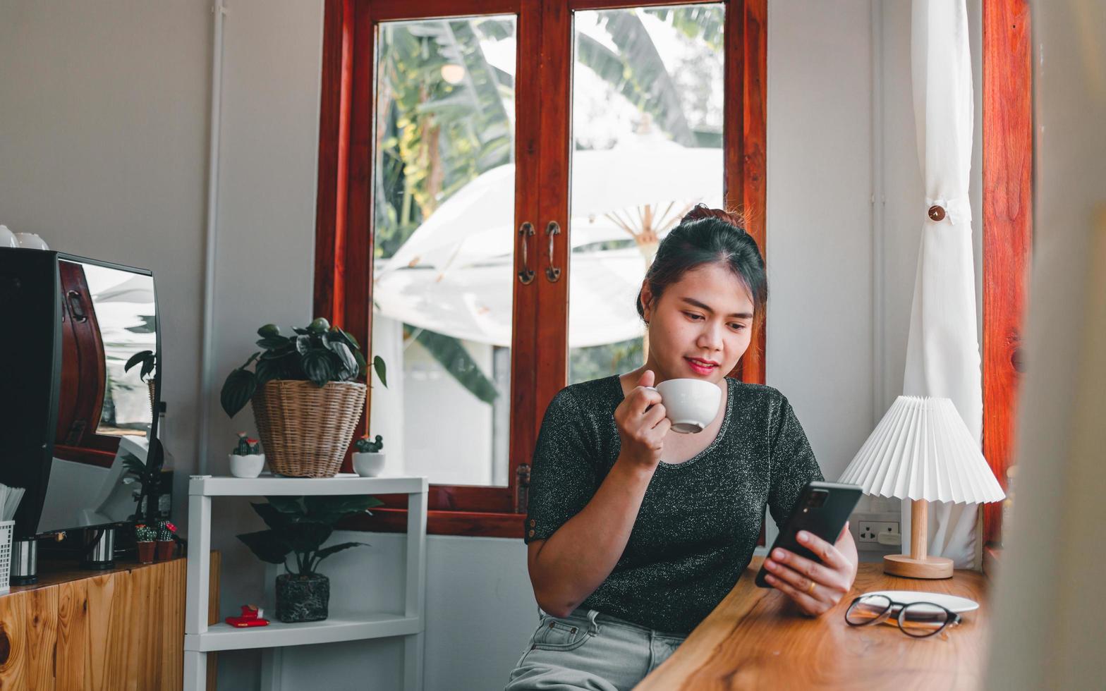 Asian woman with a beautiful smile watching on mobile phone during rest in a coffee shop, happy Thailand female sit at wooden bar counter drinking coffee relaxing in cafe during free time photo