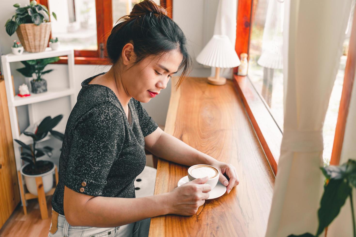 Beautiful Asian woman sits down at the bar counter inside a window coffee shop holding a cup of coffee, smiling relaxed in a cafe photo