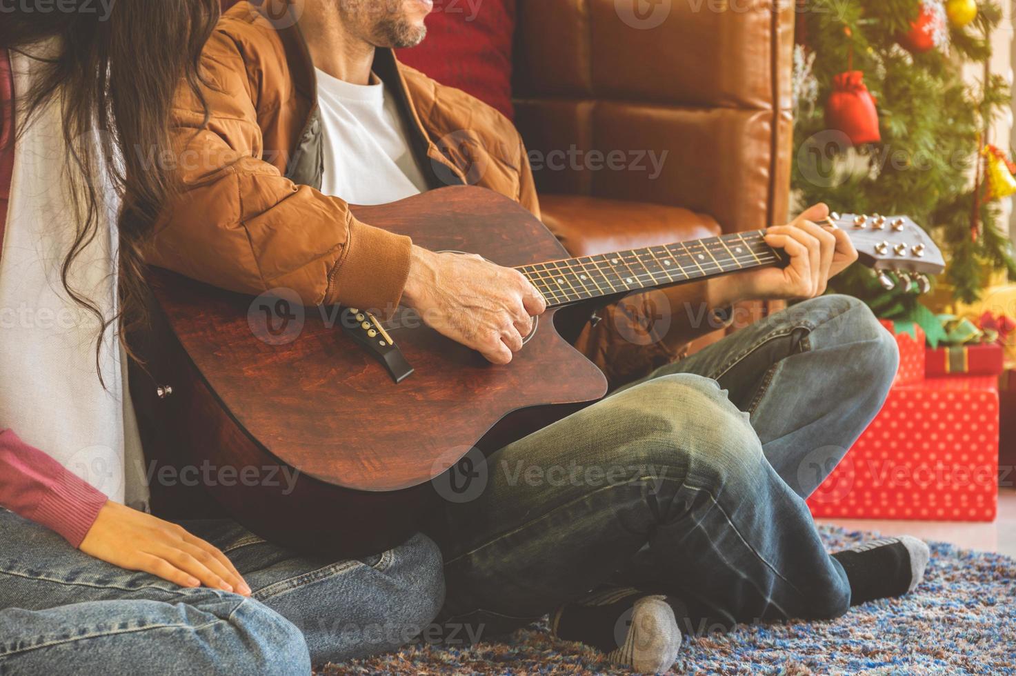 Happy cheerful couple enjoying great time in living room with guitar in Christmas holidays. Man playing guitar and singing for woman on carpet floor photo