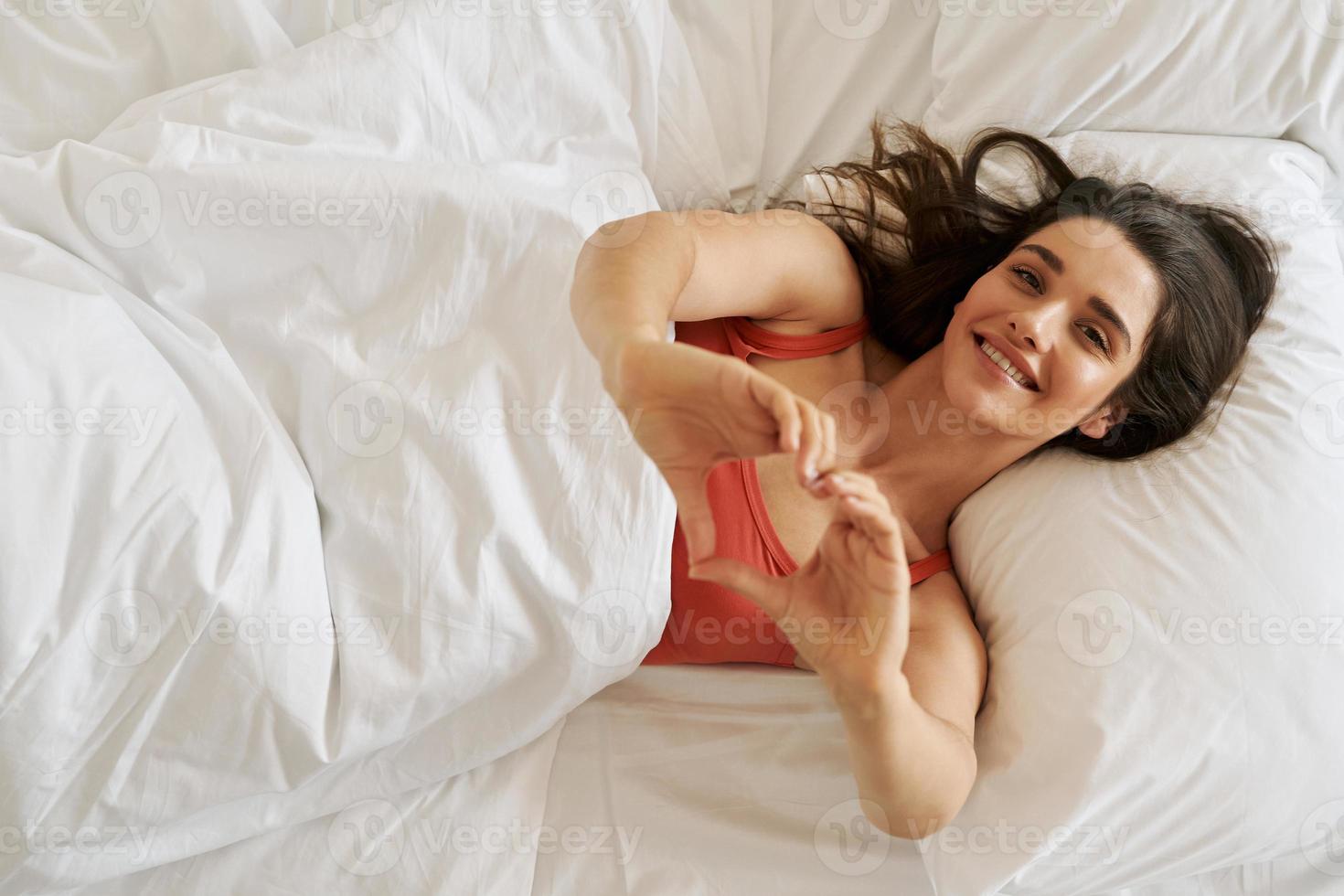 Top view of beautiful young woman gesturing heart shape and smiling while lying in bed photo