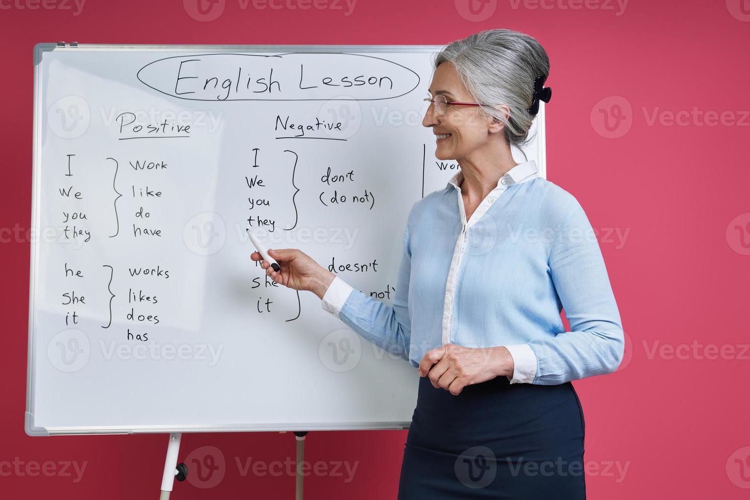 Senior woman teaching English language while standing near the whiteboard against pink background photo