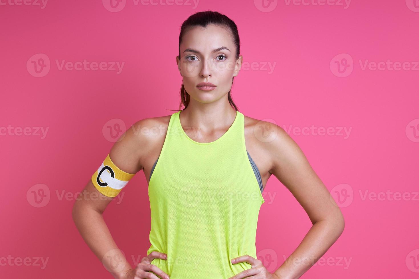 Confident woman in sports clothing wearing captain band and standing against pink background photo