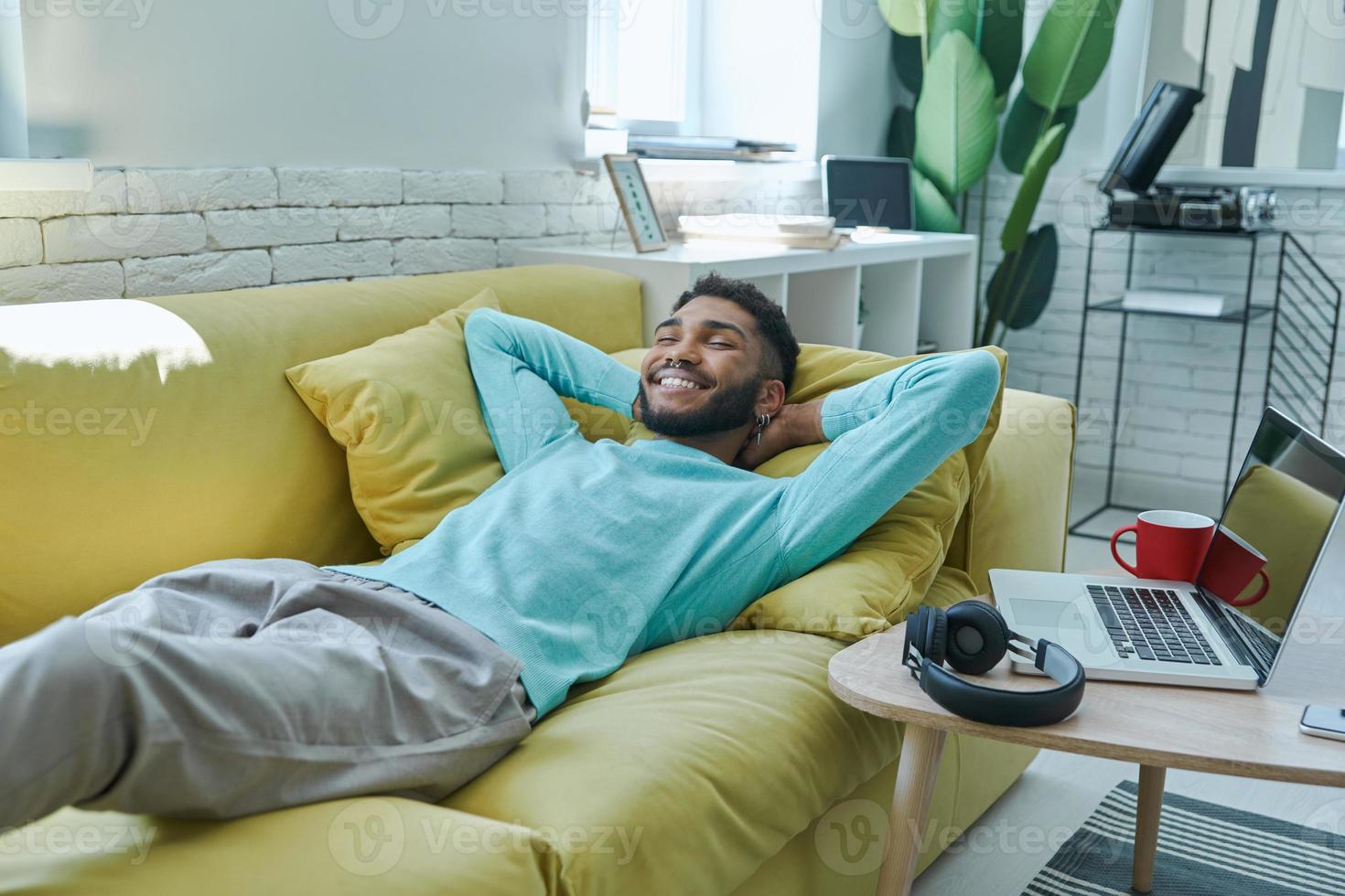 Happy African man holding hand behind head while relaxing on the couch at home photo