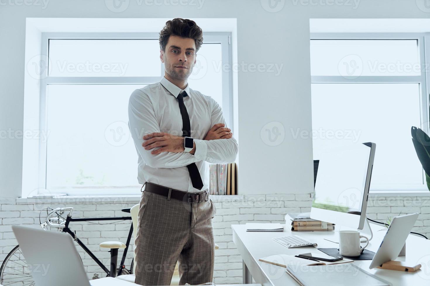 hombre guapo con camisa y corbata mirando a la cámara mientras está de pie cerca de su lugar de trabajo en la oficina foto