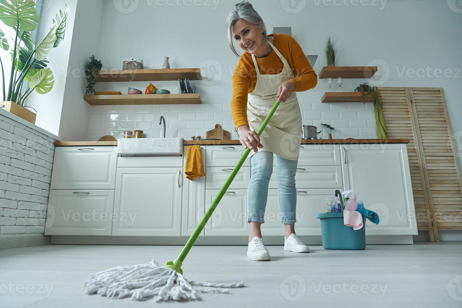 toda la longitud de la mujer mayor que limpia el piso con un trapeador mientras está de pie en la cocina doméstica foto