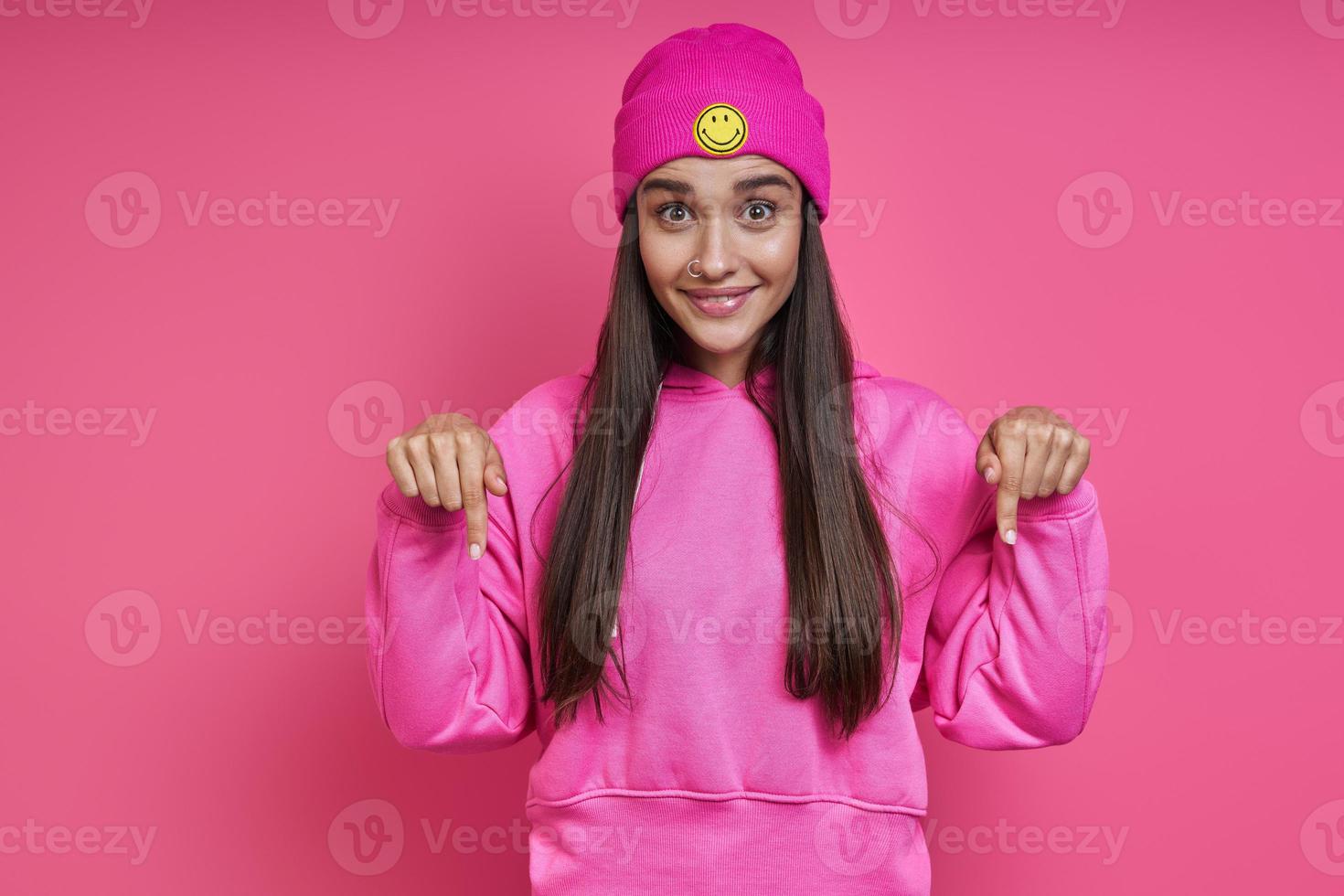 Beautiful young woman in funky hat pointing down and smiling against pink background photo