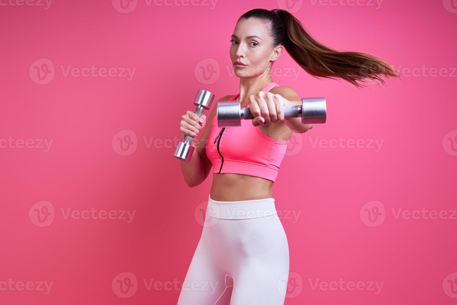 mujer joven confiada en ropa deportiva haciendo ejercicio con pesas contra fondo rosa foto