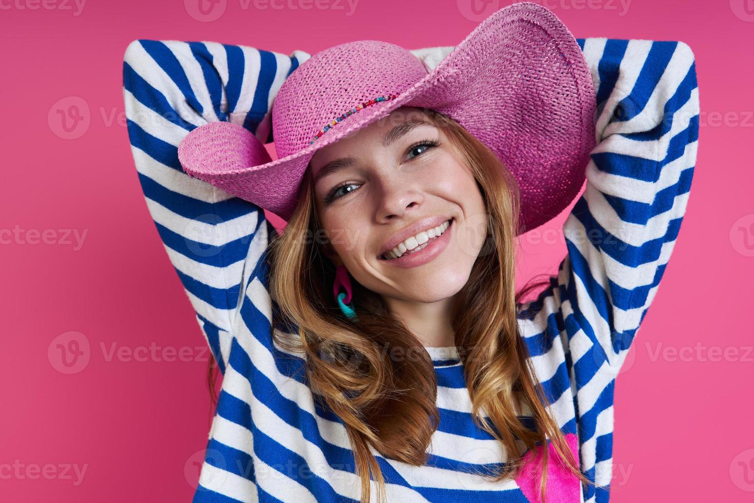 Beautiful woman in pink hat holding hands behind head while standing against colored background photo