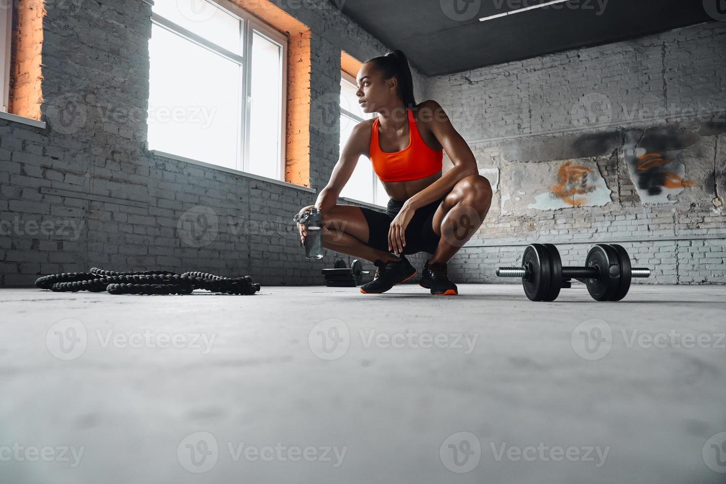 Beautiful young African woman holding bottle with water while taking a break in gym photo