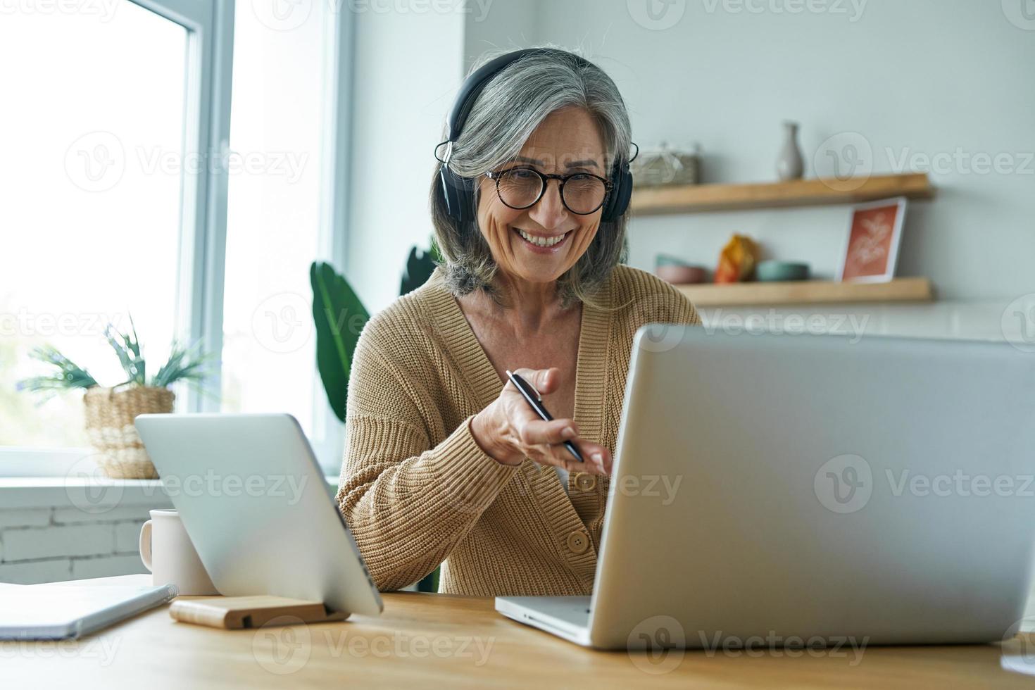 Happy senior woman using laptop and gesturing while sitting at the desk in the kitchen photo
