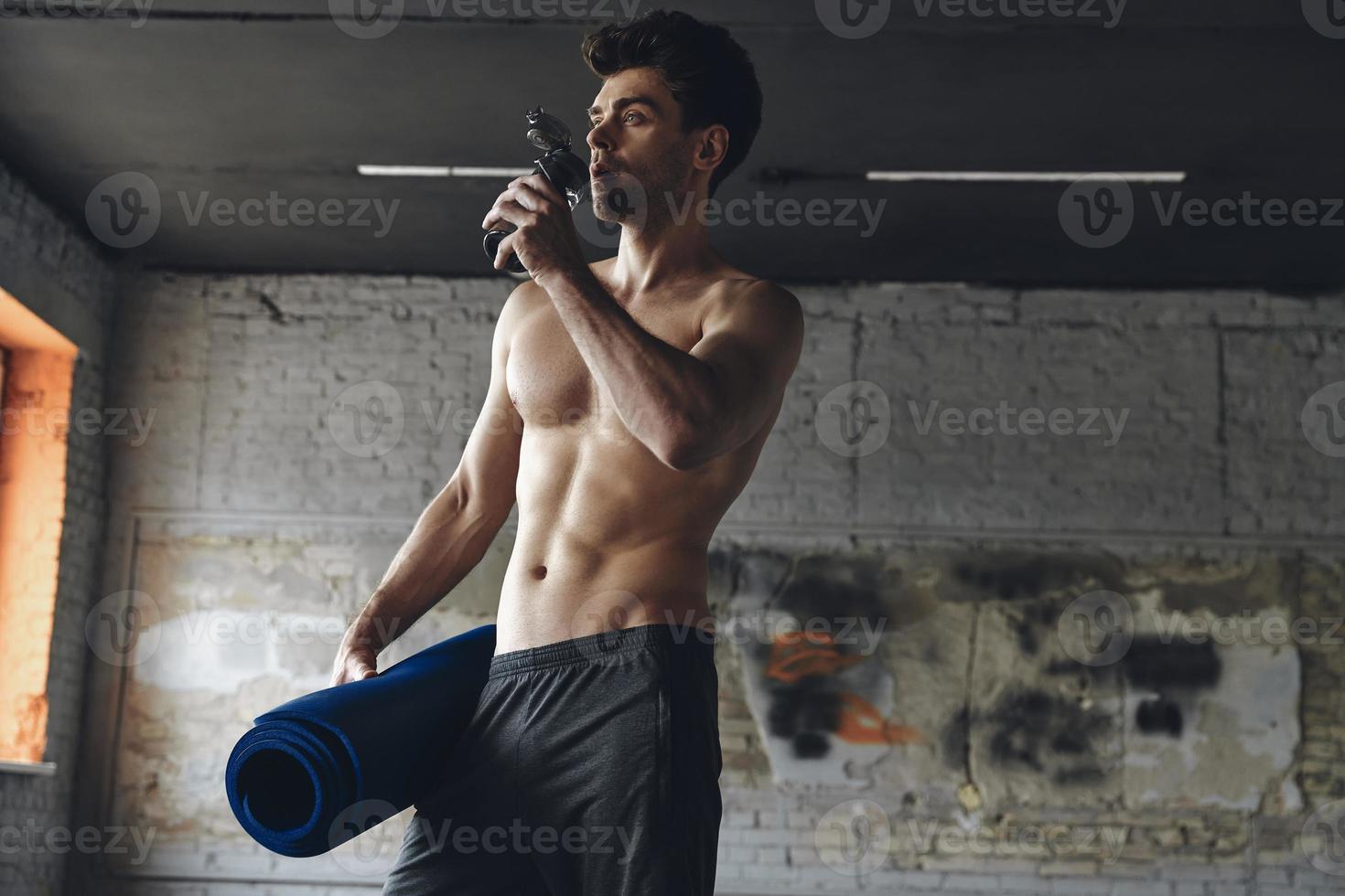 Confident young man carrying exercise mat and drinking water while standing in gym photo