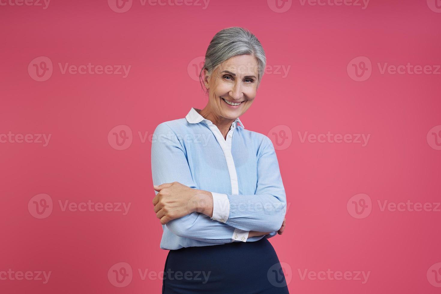 Confident senior woman keeping arms crossed while standing against pink background photo